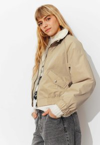 Veste courte beige avec un col texturé et duveteux, des poignets élastiques et une fermeture éclair à l'avant ; portée par-dessus un pull en tricot et un jean gris.