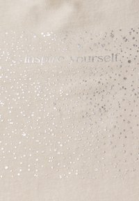 Béžová látka s vroubkovanou strukturou, na které se třpytí stříbrné jiskřičky různých velikostí a text "inspiruj se" v decentním písmu.