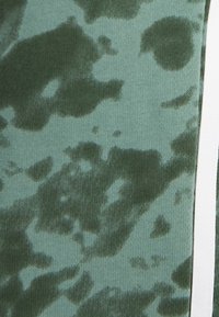 Zelená látka s vzorem a texturovaným povrchem, na níž jsou tmavé a světlé zelené skvrny. Po straně je kontrastní bílý pruh.