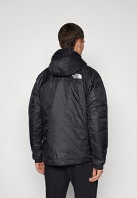 Zwarte geïsoleerde jas met een afneembare capuchon, voorzien van een gestikte textuur, elastische manchetten en een wit logo op de bovenrug.