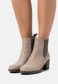 Bottines pour femmes en daim beige avec talons bas carrés et panneaux latéraux élastiques foncés, portées sur jambes nues.