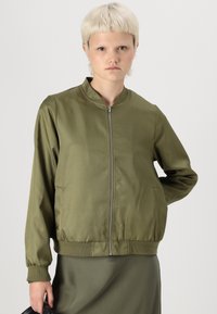 Giacca bomber verde oliva con colletto rialzato, zip frontale e polsini a costina. Texture liscia e lucida, con vestibilità leggermente ampia.