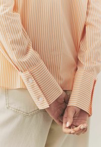Chemise à manches longues avec rayures horizontales orange et blanches, poignets boutonnés, coupe décontractée, associée à un pantalon de couleur claire.