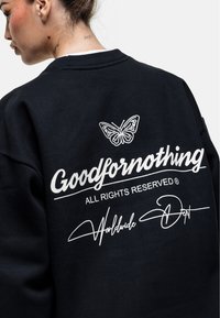 Felpa nera con grafica di una farfalla bianca e il testo "Goodfornothing" sul retro. Texture morbida, design a girocollo e vestibilità ampia.