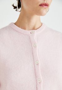 Απαλή ροζ ζακέτα από μαλακό ύφασμα, με στρογγυλή λαιμόκοψη, ραφές με λάστιχο και πέντε άσπρα κουμπιά μπροστά.