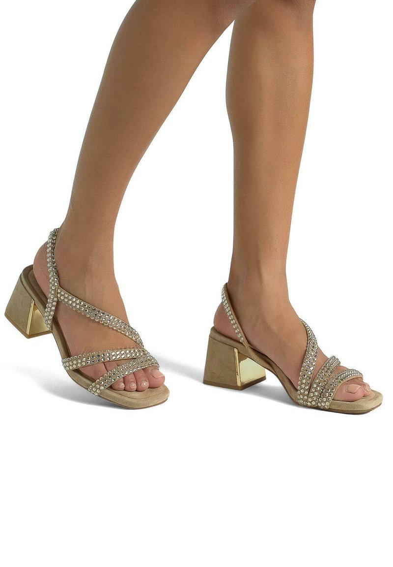 Beige Riemchensandalen mit dekorativen Strasssteinen, einem niedrigen Blockabsatz und metallischen Akzenten am Absatz, die offene Zehen und gekreuzte Riemen zeigen.