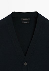 Svart bomullskardigan med V-ringning, ribbad textur och en enda knappstängning. Etiketter anger märke och skötselanvisningar.