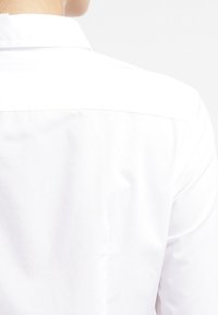 Camicia bianca con un tessuto liscio e strutturato. La parte posteriore presenta una cucitura a yoke, e il colletto è piegato ordinatamente piatto.