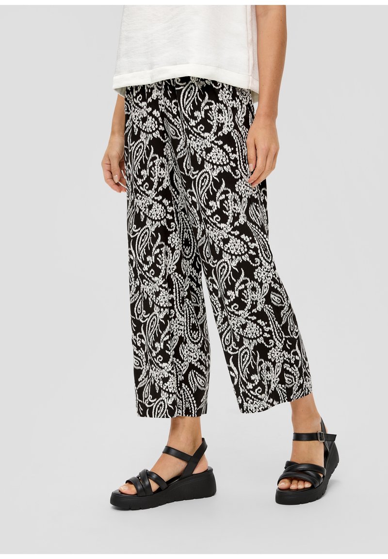 Personne portant un pantalon ample à motif floral noir et blanc et des sandales compensées noires avec des brides à la cheville.