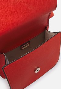 Rød lædertaske med en struktureret overflade, guldfarvet tryklås og beige interiør med et mærke-tag. Kompakt rektangulært design.