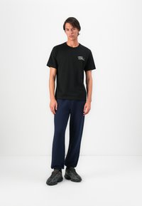 T-shirt en coton noir avec un graphique sur le devant, associé à un pantalon de survêtement bleu marine. Le mannequin porte des baskets grises avec des détails texturés. Design simple, coupe décontractée.