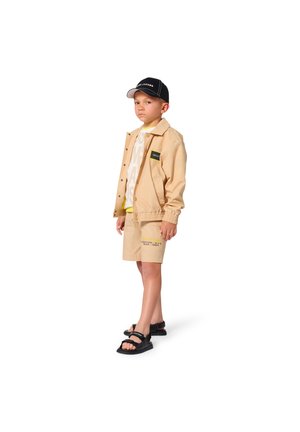 Ragazzo con giacca beige e pantaloncini, maglietta bianca, cappello nero e sandali neri, in piedi e che guarda avanti su uno sfondo bianco.