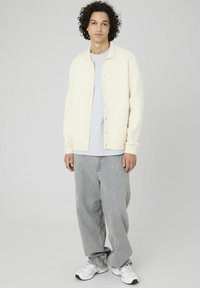 Un gilet en tricot crème clair avec des boutons, accompagné d'un T-shirt gris et d'un pantalon gris ample. Les chaussures sont des baskets blanches.