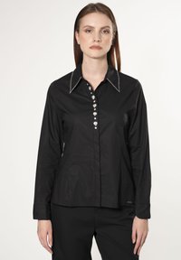 Camicia nera a maniche lunghe con colletto, caratterizzata da una fila di bottoni decorativi e cuciture bianche lungo il bordo del colletto.