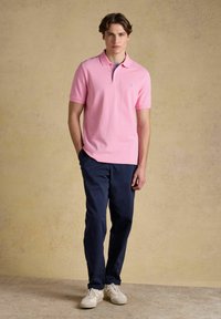 Jeune homme portant un polo rose, un pantalon bleu marine et des baskets blanches, debout devant un fond beige uni.