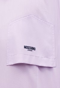 Τσέπη από ύφασμα λεβάντας με εξαγωνικό σχήμα και ορατή ραφή, κεντημένη με μαύρο κείμενο "Moschino Jeans" στο κέντρο.