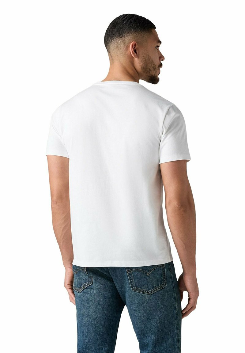 Mann mit kurzen Haaren, der ein schlichtes weißes kurzärmeliges T-Shirt und blaue Jeans trägt, von hinten gesehen, schaut nach rechts.