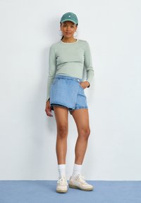 Femme portant un haut à manches longues vert clair, un short en jean, une casquette turquoise, des chaussettes blanches et des baskets beiges, se tenant devant un mur blanc et un sol bleu.