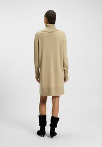 Beiges Strickkleid bis zum Knie mit hohem geripptem Kragen, langen Ärmeln und gerippten Saumdetails. Das Model trägt schwarze lässige Stiefel.