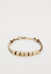 Bracelet en chaîne dorée composé de maillons carrés avec des lettres gravées formant "LACOSTE". Texture lisse avec une finition polie. Fermoir ajustable.