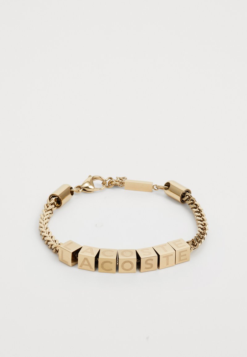 Bracelet en chaîne dorée composé de maillons carrés avec des lettres gravées formant "LACOSTE". Texture lisse avec une finition polie. Fermoir ajustable.