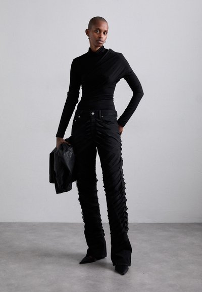 Haut noir ajusté à manches longues et pantalon taille haute avec une texture froncée et des jambes évasées. Le mannequin tient une veste noire.