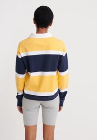 Polo a maniche lunghe a righe gialle, blu navy e bianche con colletto. Pantaloni corti grigi indossati sotto. Tessuto di cotone liscio con una vestibilità rilassata.