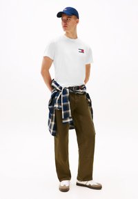 Mand iført hvid Tommy Jeans T-shirt, marineblå kasket, olivengrønne bukser, hvide sneakers og blå ternet skjorte bundet omkring taljen, kigger mod højre.