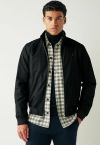 Sort lynlåsjakke lagdelt over en beige og navy ternet skjorte med en mock neck sweater under. Glat stof, afslappet pasform.