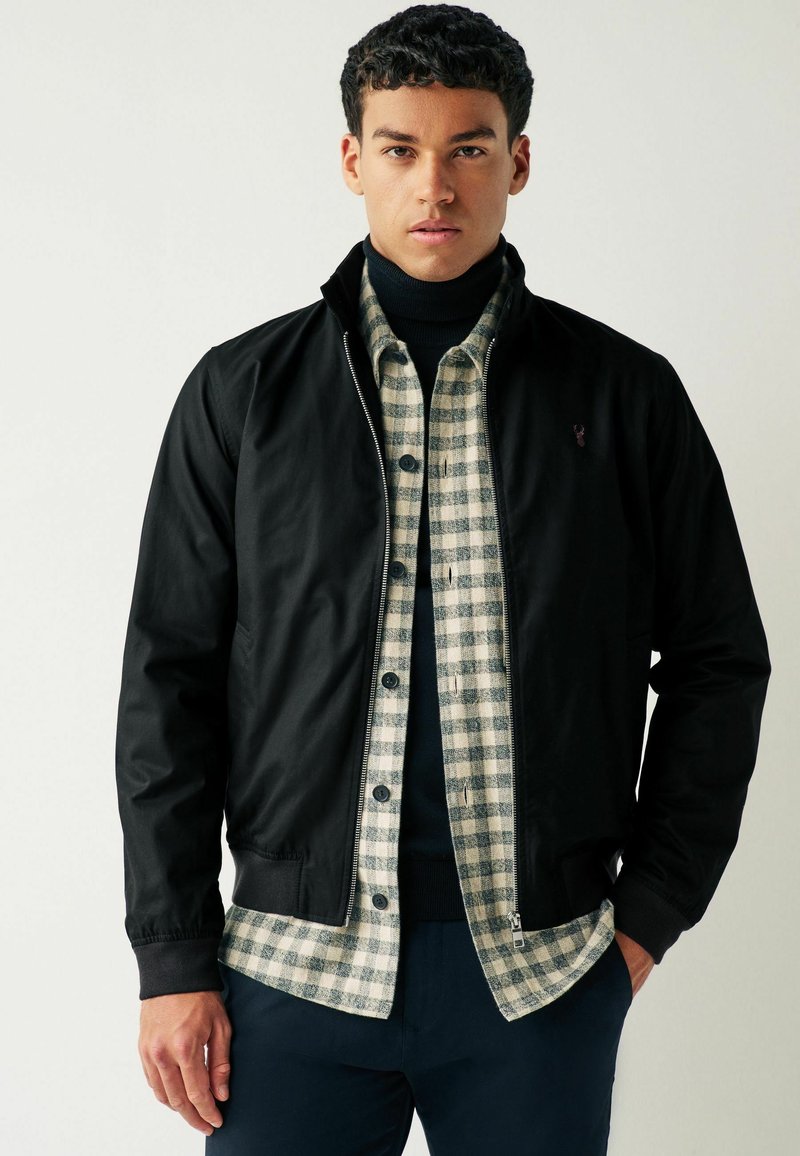 Giacca nera con zip indossata sopra una camicia a quadri beige e blu navy, con un maglione a collo alto sotto. Tessuto morbido, vestibilità casual.