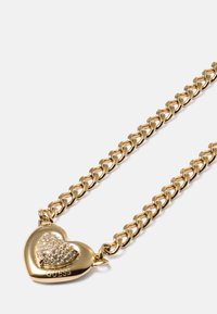Colar de corrente dourada com um pendente em forma de coração com superfície texturizada e detalhes em strass. O nome da marca "GUESS" é visível.