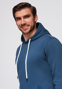 Felpa blu realizzata in un tessuto morbido, con tasca frontale e cordini bianchi. Texture liscia e design con vestibilità rilassata.