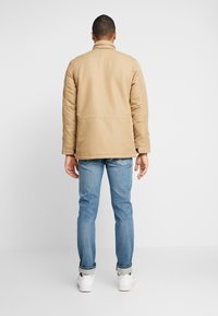 Beige jacka med krage, långa ärmar och en avslappnad passform, kombinerad med ljusblå jeans och vita sneakers, sedd bakifrån.