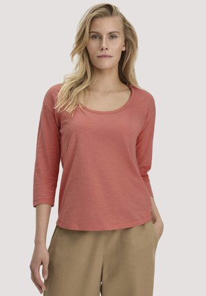 Blonde Frau trägt ein staubiges rosa langärmeliges Hemd und beige Hosen, steht mit einer Hand in der Tasche vor einem schlichten Hintergrund.