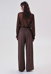 Bruine geruite wijde broek met een hoge taille, gecombineerd met een doorschijnende zwarte blouse met lange mouwen. Soepele textuur met een losse pasvorm.