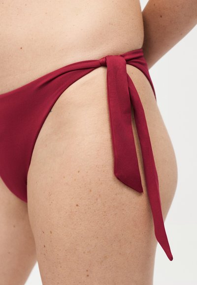 Κοντινή λήψη γοφού που φοράει μπορντό κατώτερο μέρος μαγιό δεμένο στο πλάι με κόμπο, σε φωτεινό φόντο.