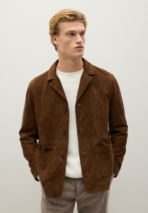 Giacca in suede marrone con colletto a punte, due tasche frontali e chiusura a bottoni, indossata sopra un pullover bianco con texture.