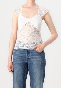 Haut en dentelle blanche avec manches courtes, présentant un design en couches et des bords festonnés, porté avec un jean en denim bleu taille haute.