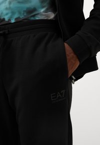Schwarze Jogginghose mit einem Kordelzugbund, ausgestattet mit einem kleinen EA7-Logo. Das Material wirkt weich und glatt mit einer lockeren Passform.
