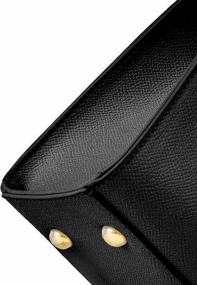 Črni teksturirani spodnji vogal usnjene torbice z dvema okroglima zlatima kovinskima zatičema na belem ozadju.