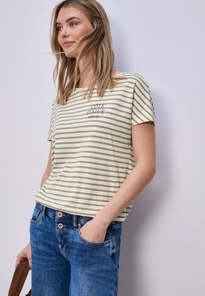 Jeune femme portant un T-shirt beige et blanc rayé avec le texte "COTE D'AZUR" et un jean bleu, debout les mains dans les poches.
