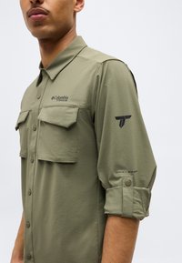 Άνδρας που φοράει πουκάμισο Columbia Titanium σε ελιά πράσινο, με κουμπιά, μανίκια που έχουν σηκωθεί και τσέπες στο στήθος, μπροστά σε απλό φόντο.