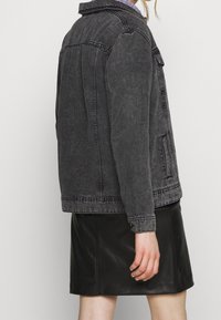 Giacca in denim nera con un design retrò, caratterizzata da un colletto a punta, chiusura con bottoni e due tasche laterali. Indossata sopra una gonna di pelle nera.