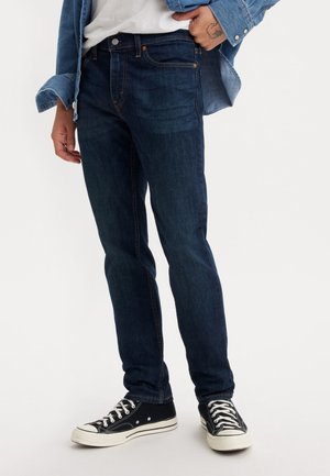 Persoon draagt donkerblauwe slim-fit jeans, zwart-witte sneakers, wit overhemd en gedeeltelijk open spijkerjack met één hand in de zak.