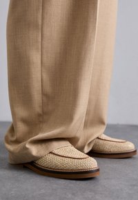 Vevede beige loafers med brune detaljer og gummisåle, i kombinasjon med løse, lysebrune bukser som dekker skoens overdel.