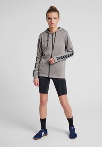 Grauer Reißverschluss-Hoodie mit schwarzen Chevron-Akzenten an den Ärmeln, kombiniert mit schwarzen Radshorts und blauen Sportschuhen. Einfaches, funktionales Design.
