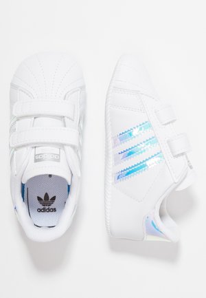 Par hvide Adidas småbørnssko med iriserende tre striber og dobbelte velcrobånd på ensfarvet baggrund.