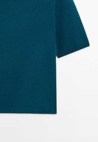 Pull tricot à manches courtes bleu sarcelle avec poignets et ourlet côtelés sur fond blanc, montrant uniquement l'épaule et la manche droite.