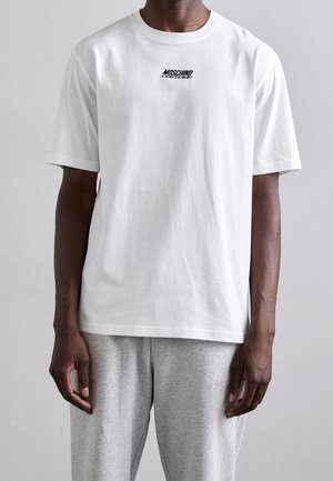 Homme portant un t-shirt blanc Moschino Couture et un pantalon gris clair, debout devant un fond gris clair uni.