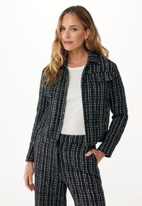 Giacca in tessuto a scacchi bianco e nero con colletto, tasche frontali e un design leggermente corto. Abbinata a una camicia bianca.